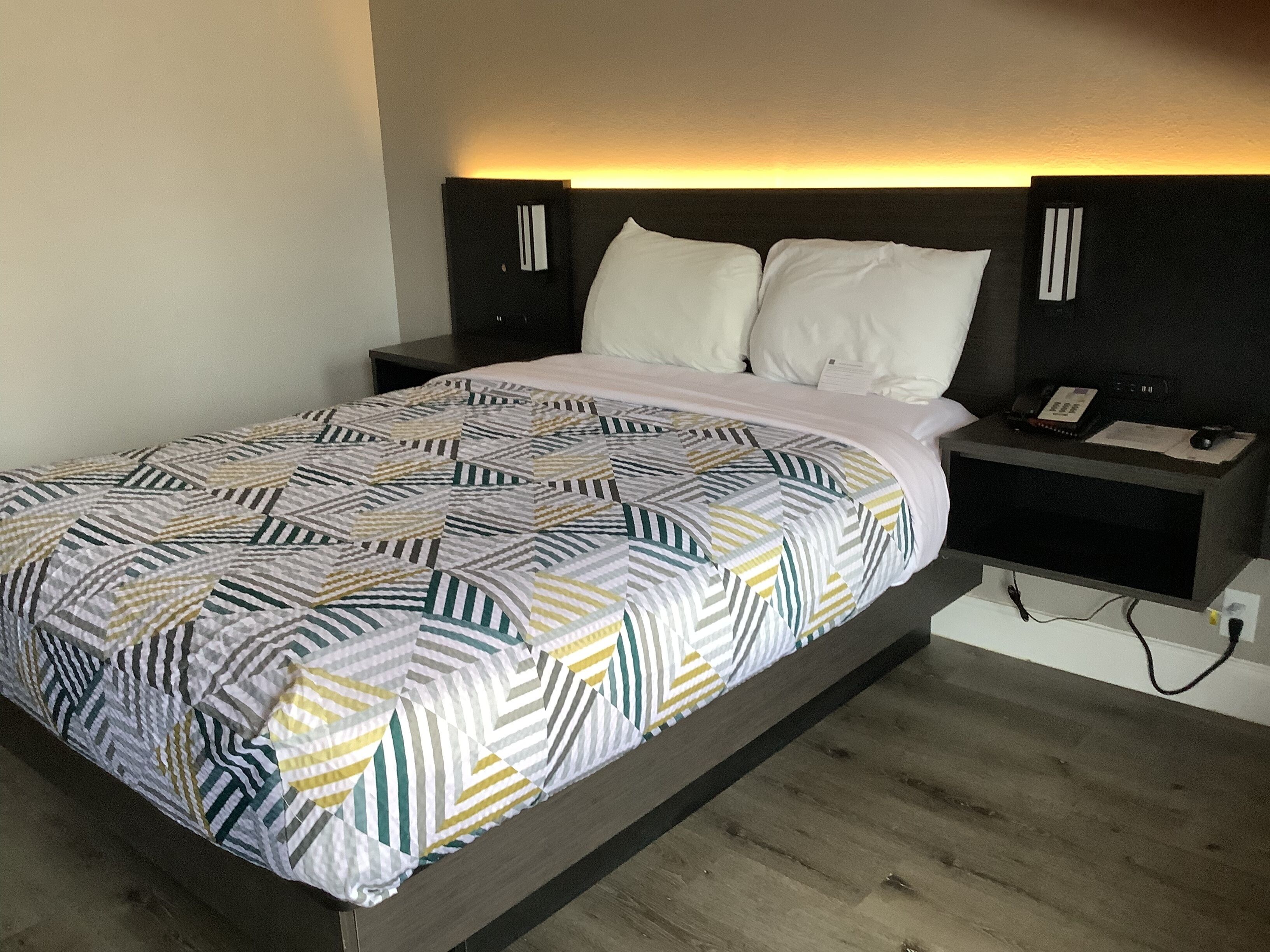 A modern bedroom with a geometric-patterned bedspread, a dark headboard, and a bedside table with a phone and papers.