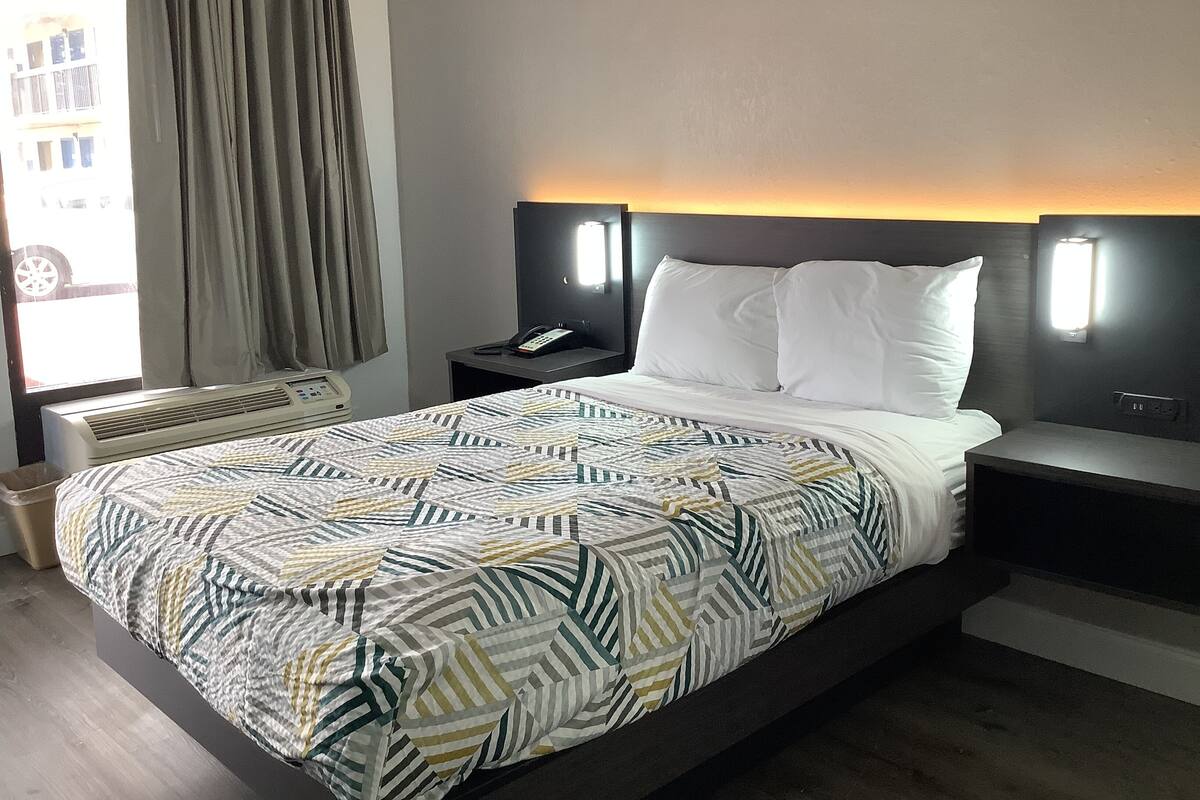 Habitación de hotel con cama, mesita de noche con teléfono, lámpara de pared y ventana con cortinas.