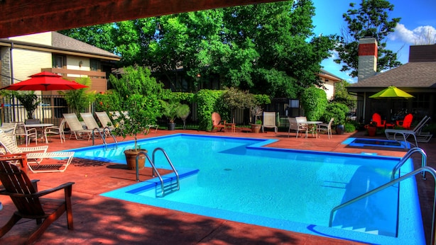 A rectangular swimming pool with a blue edge, surrounded by lounge chairs and umbrellas, with trees and a building in the background. - The Inn at Tallgrass (Wichita)