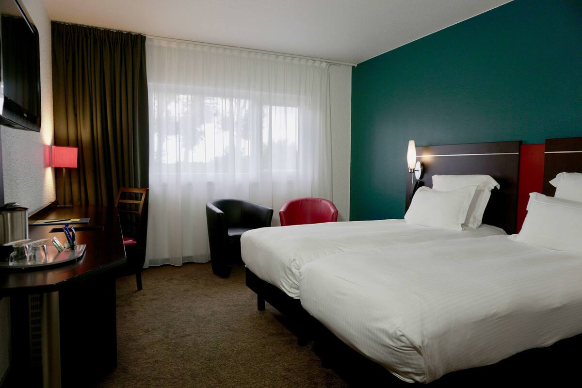Une chambre d’hôtel avec un grand lit, un bureau avec une lampe, une chaise et une fenêtre avec des rideaux.
