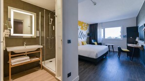 A modern hotel room with a bathroom featuring a glass-enclosed shower, a sink, and a mirror. The room includes a bed, a desk, a chair, and a TV.