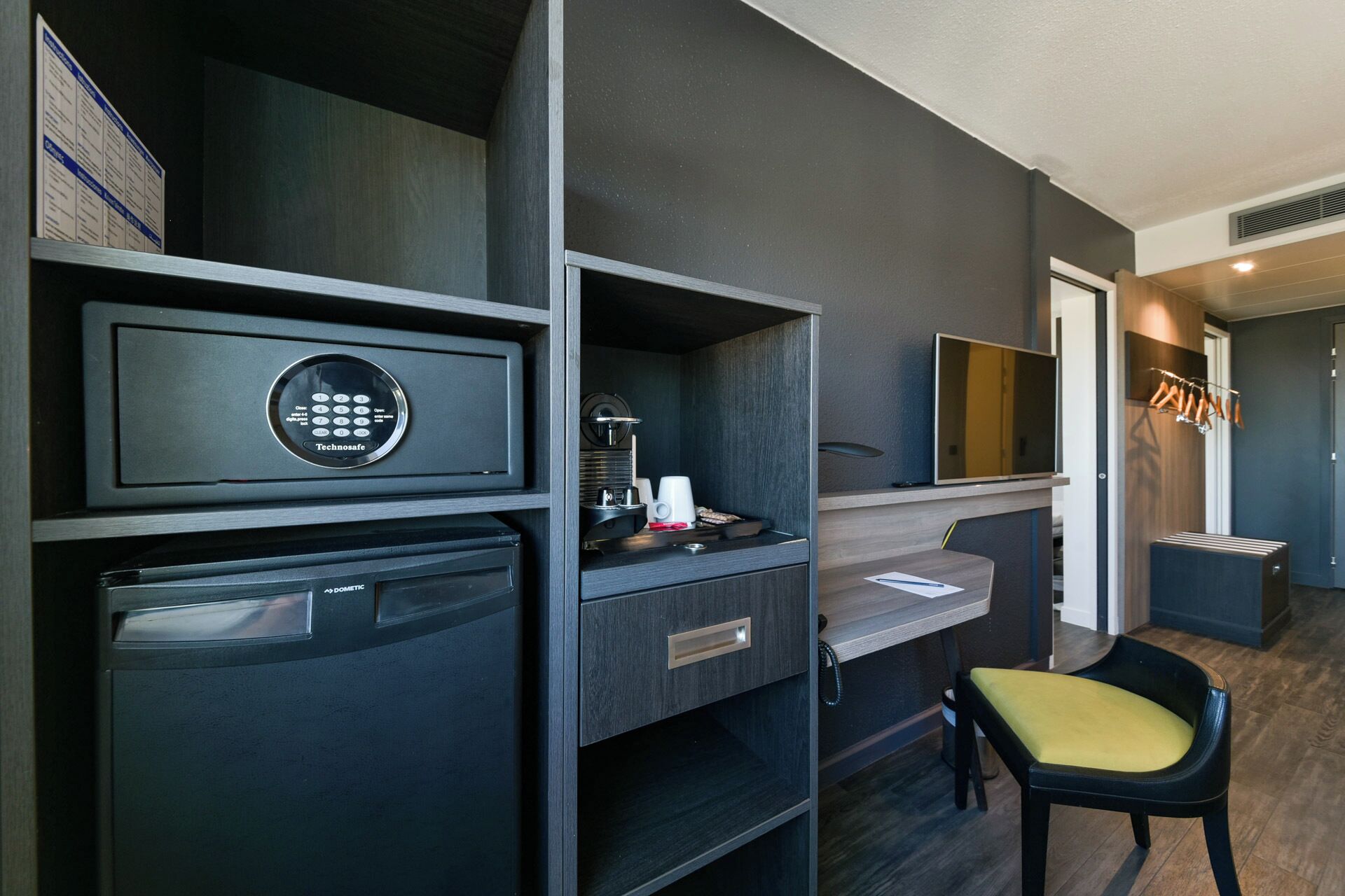 A compact hotel room with a minibar, a desk with a coffee machine, a chair, and a TV mounted on the wall.