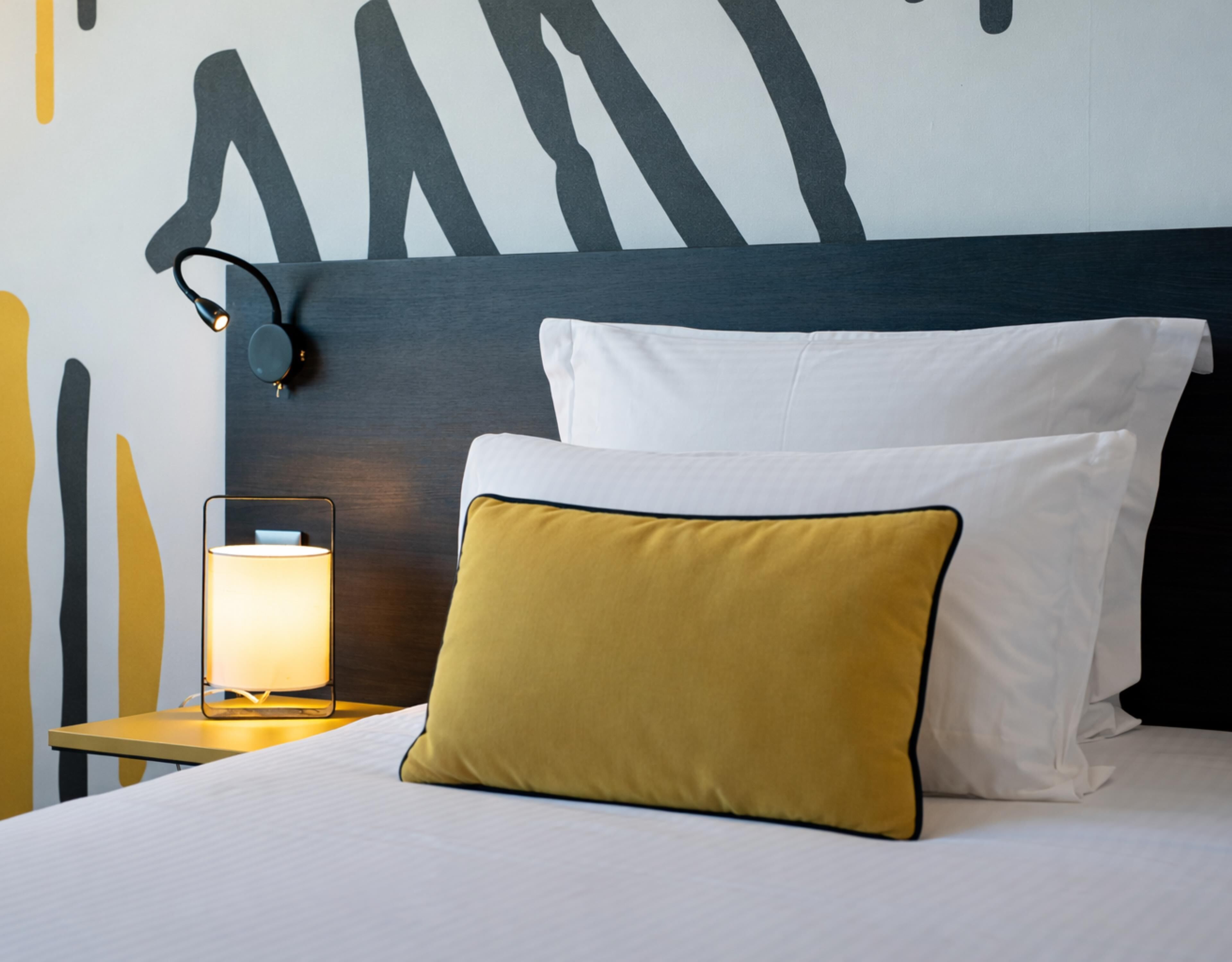 A neatly made bed with white and mustard pillows, a bedside lamp, and a modern wall design.