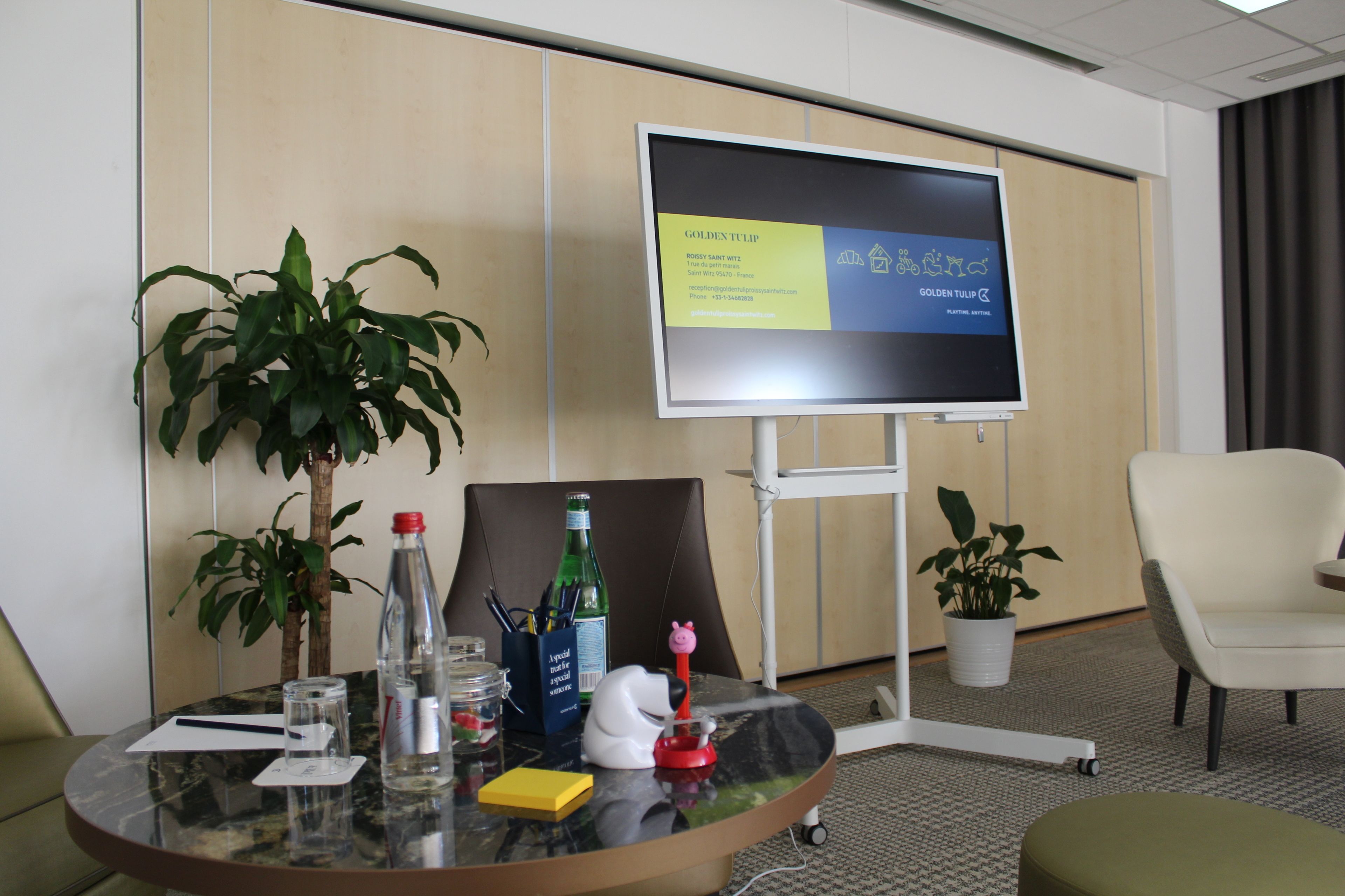 A conference room with a large screen displaying a presentation, a glass table with various items, two potted plants, and a white chair.