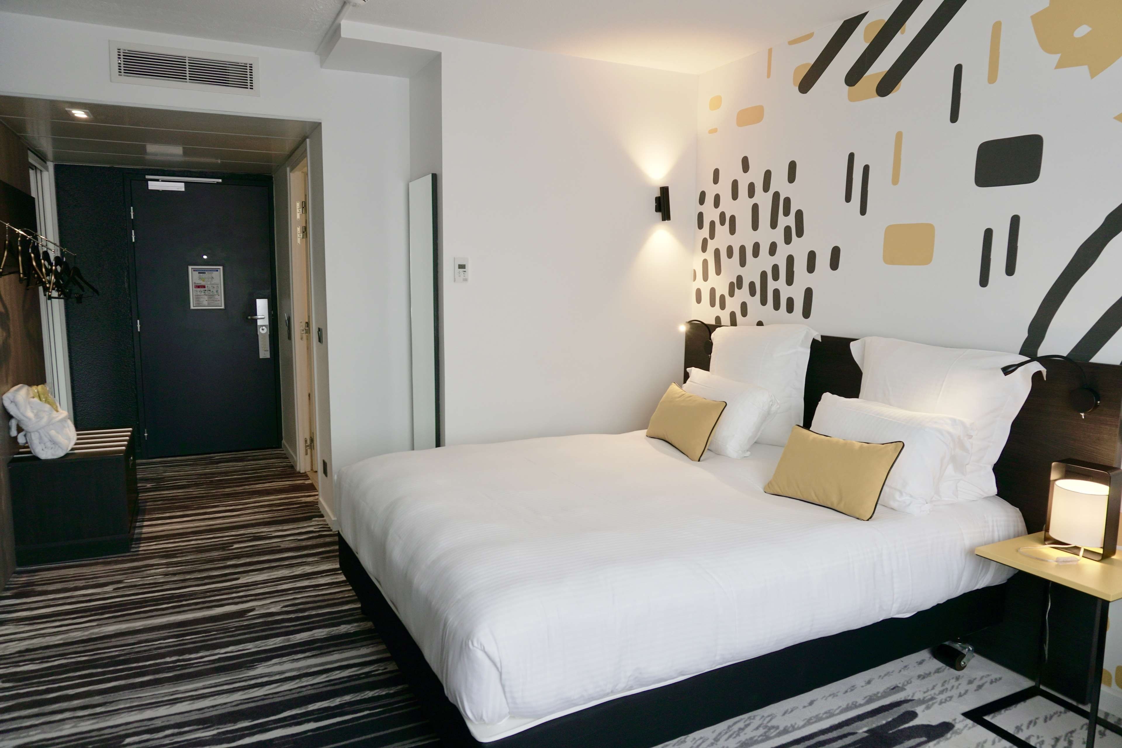 A hotel room with a large bed, bedside tables, a wall-mounted lamp, and a door leading to a hallway.