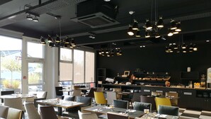 A restaurant with multiple dining tables and chairs, a kitchen area with a buffet, and large windows allowing natural light.