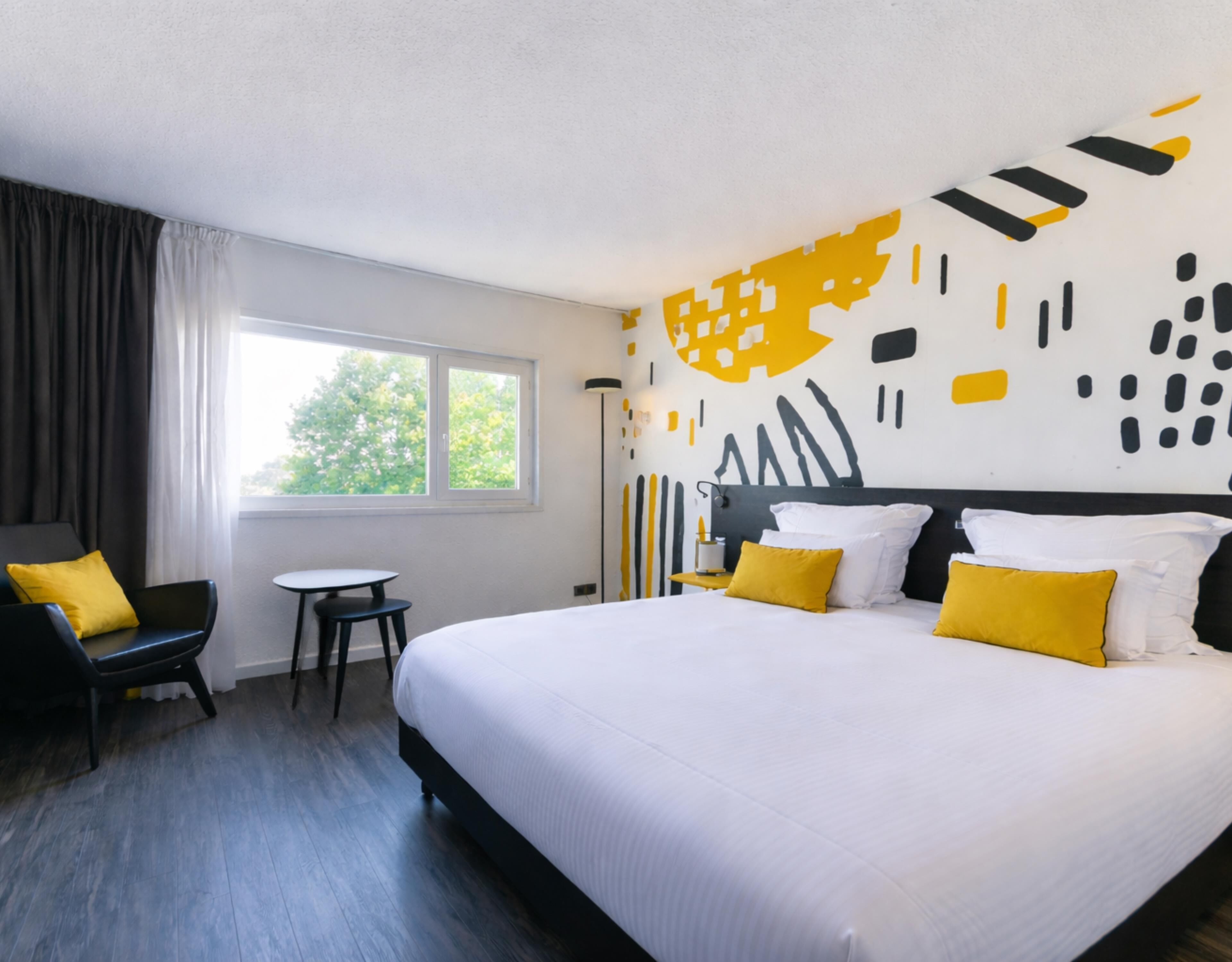 Hotellihuone, jossa on suuri sänky, pieni pöytä, tuoli ja ikkuna, jossa on verhot.