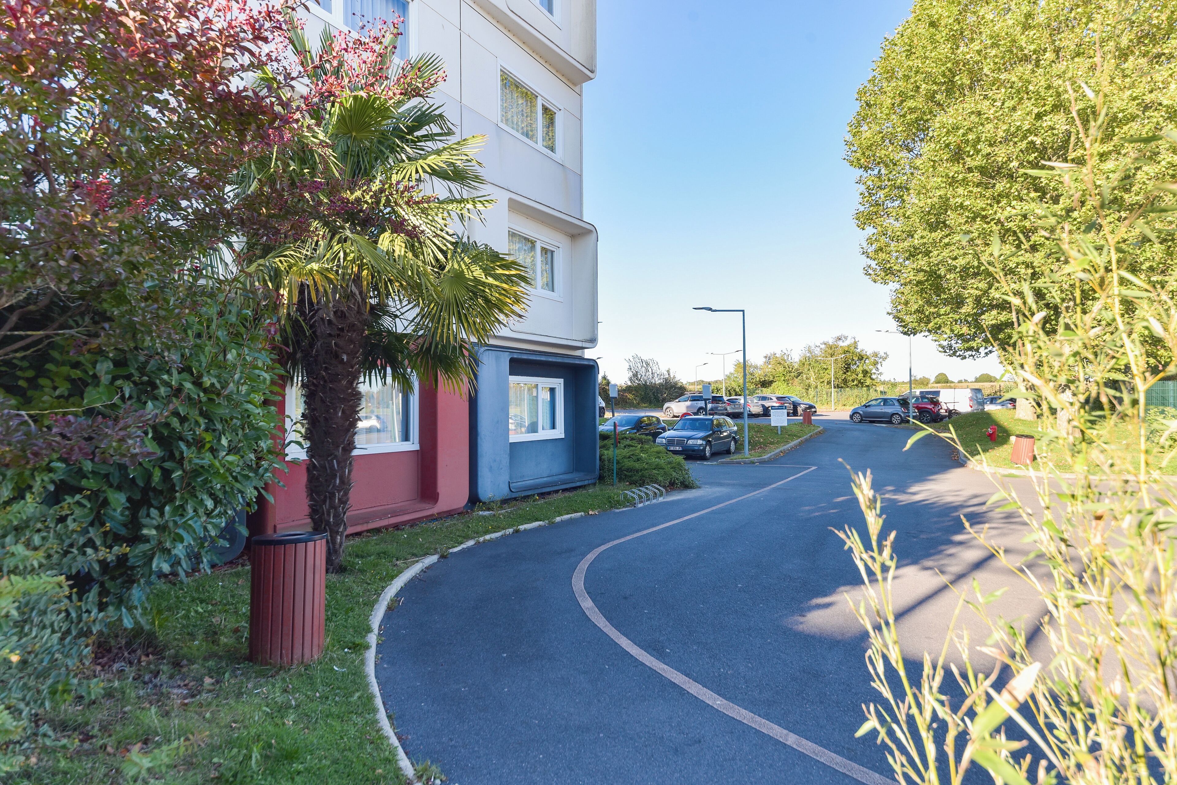 Une route sinueuse mène à un bâtiment à la façade rouge et bleue, entouré de verdure et de voitures garées.