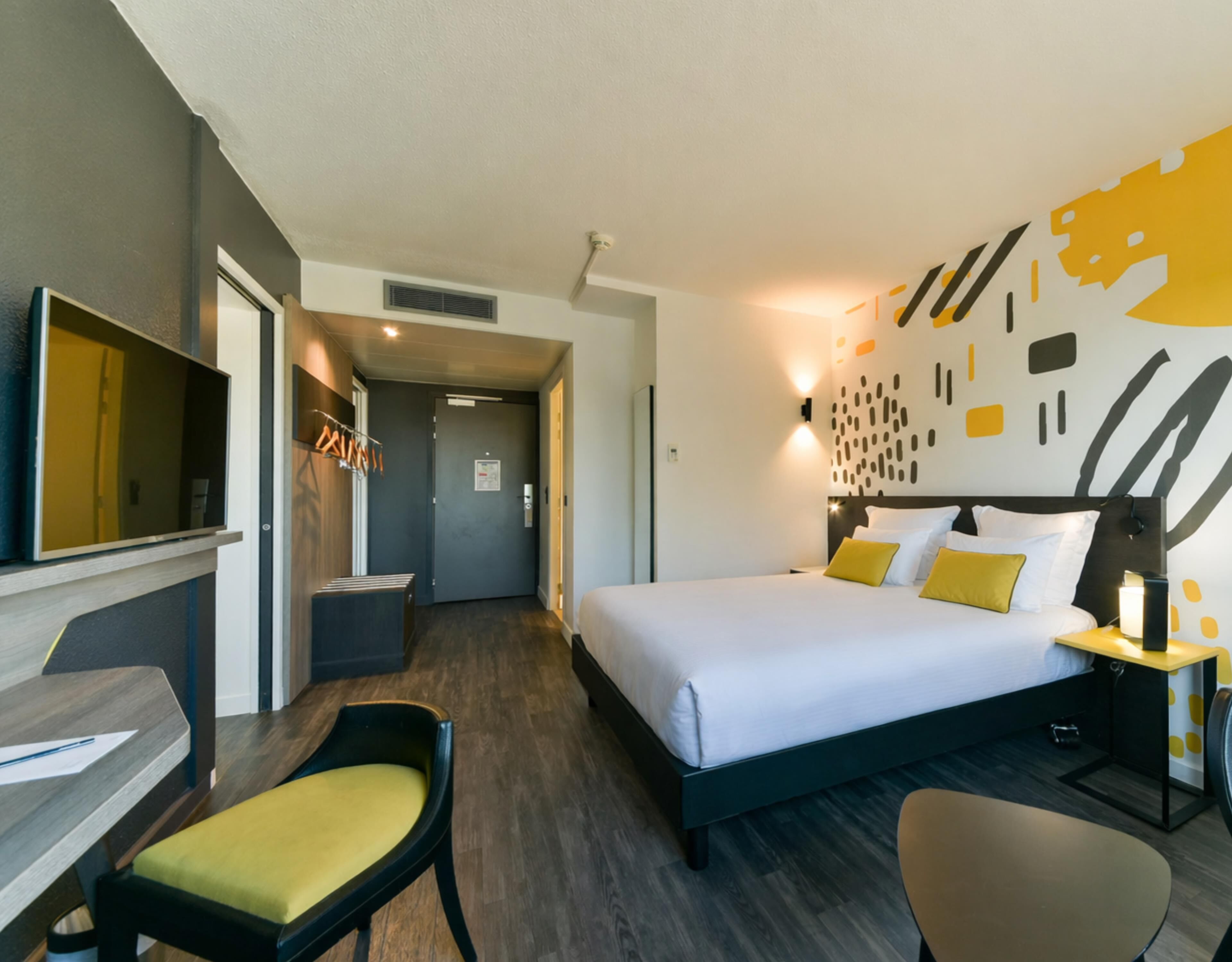 Ein Hotelzimmer mit Bett, Schreibtisch, Stuhl, Fernseher und einer Wand mit abstrakter Kunst.
