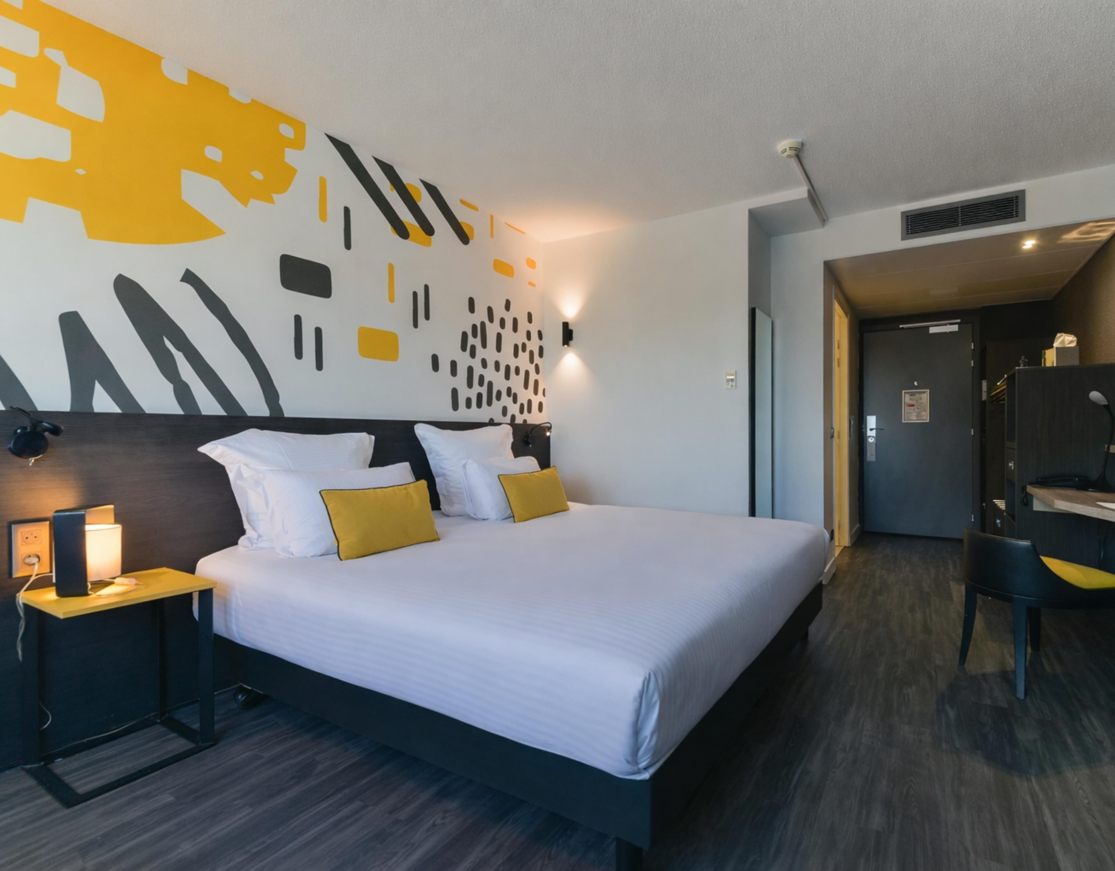 Ein Hotelzimmer mit einem großen Bett, einem Nachttisch mit Lampe, einer Wand mit abstrakter Kunst und einer Tür zu einem Badezimmer.