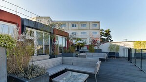 A rooftop patio with outdoor seating, planters, and a view of a multi-story building in the background.