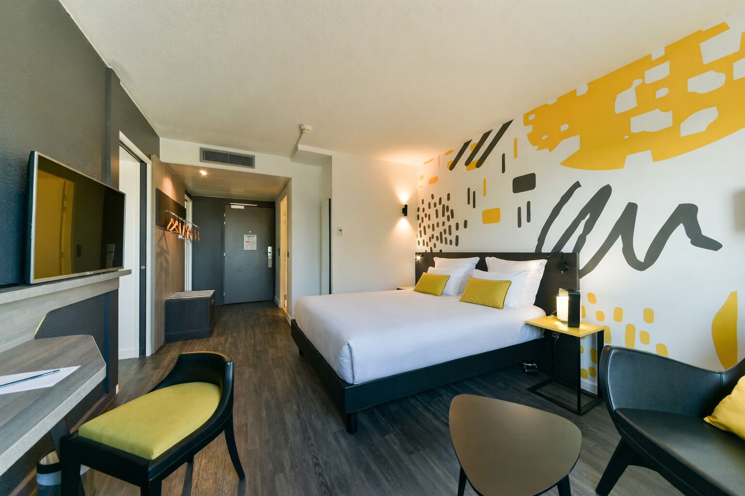 Een moderne hotelkamer met een bed, een bureau en een stoel. Er is een gang die naar een andere kamer leidt.