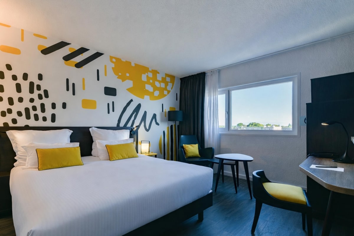 A hotel room with a large bed, a desk, a chair, a small table, and a view of the outdoors.