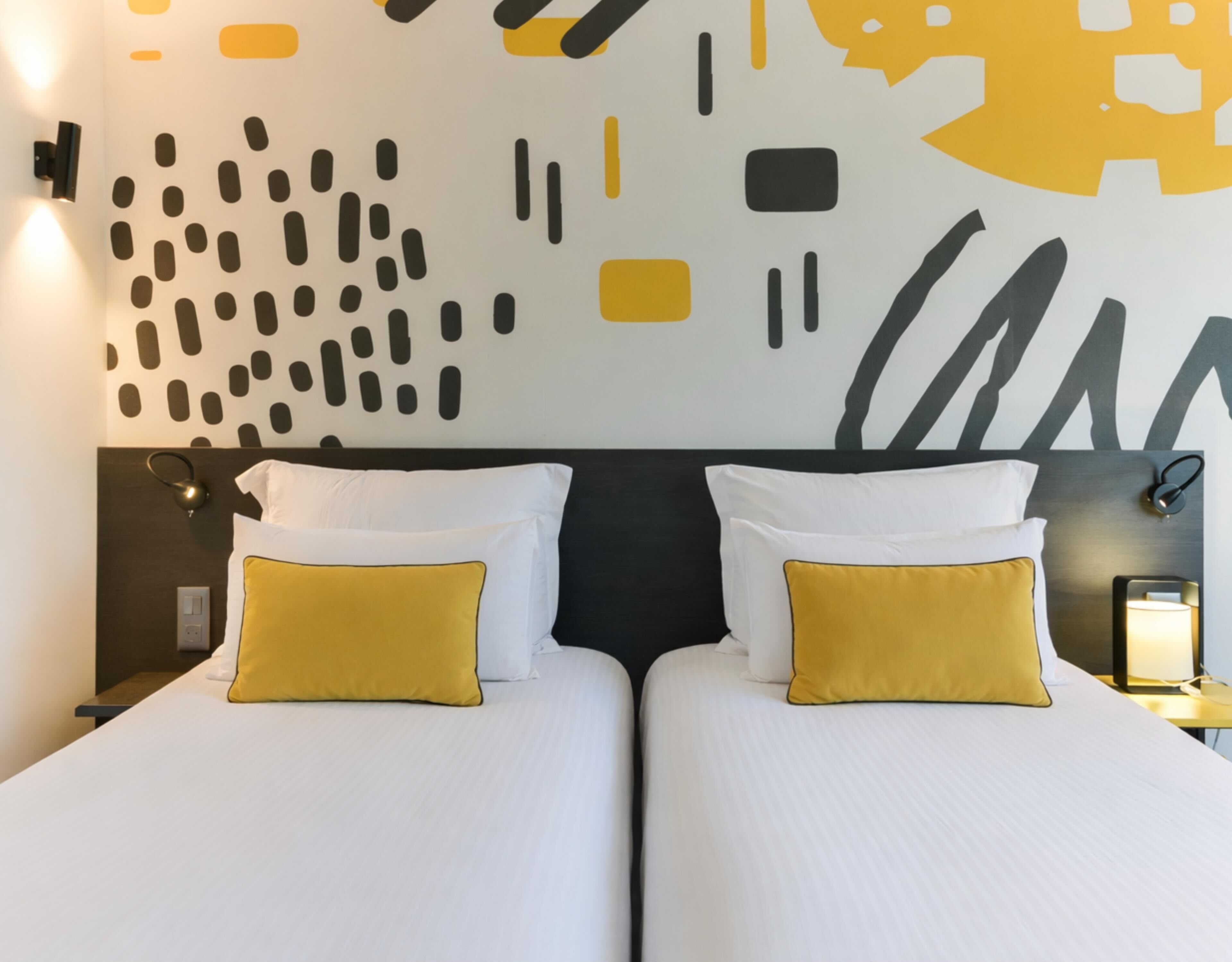 A hotel room with two beds, each with white bedding and a single yellow pillow. The room features a modern wall design with abstract patterns and a dark headboard.