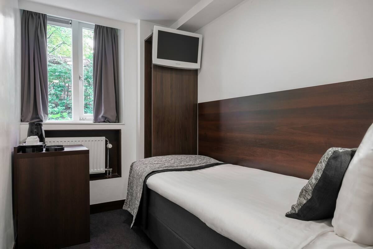 Ein Schlafzimmer mit einem Bett, einem darüber montierten Fernseher, einem Schreibtisch mit Lampe sowie einem Fenster mit Vorhängen.