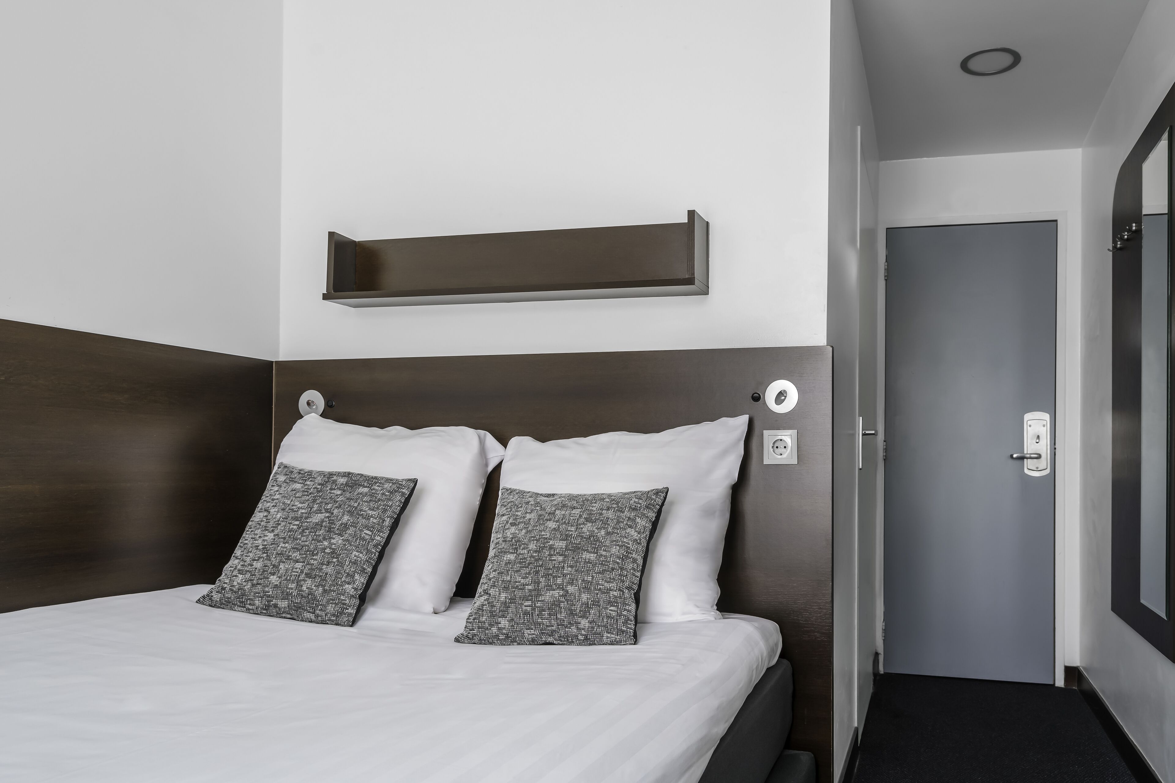Un lit bien fait, avec du linge de lit blanc et deux coussins gris à motifs, une tête de lit en bois foncé et une petite étagère au-dessus du lit.
