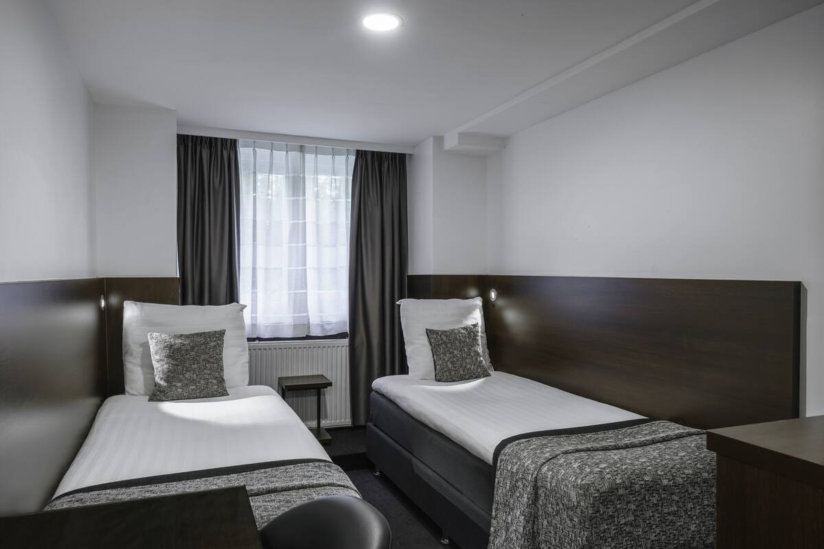 Ein Hotelzimmer mit zwei Einzelbetten, einem kleinen Tisch und einem Fenster mit Vorhängen.