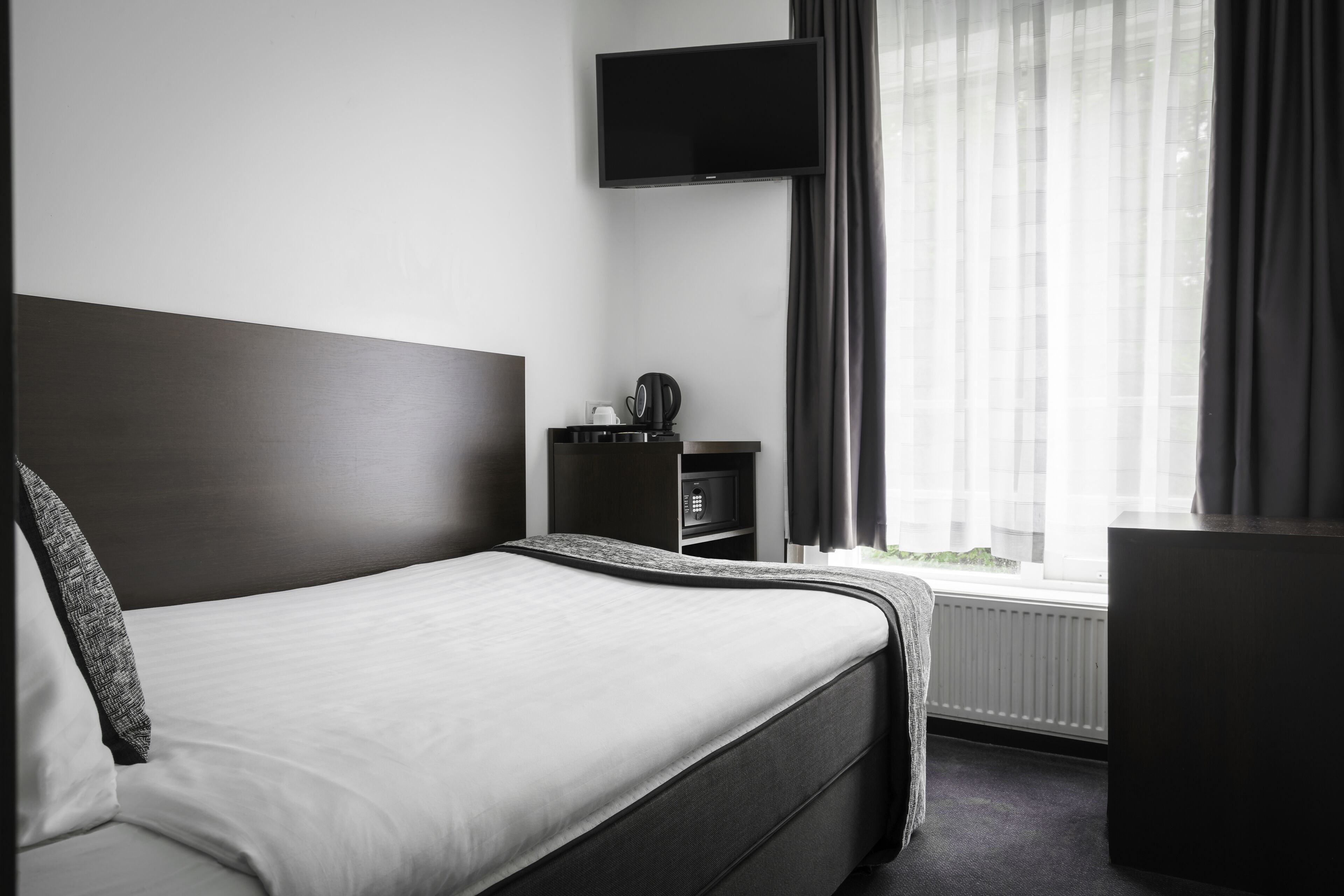Habitación de hotel moderna con una cama grande, un televisor de pantalla plana en la pared y un ventanal con cortinas translúcidas.