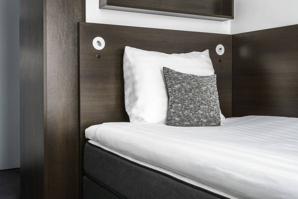 Ein modernes Hotelzimmer mit einem dunklen Holz-Kopfteil, einem weißen Bett und einem grauen Kissen mit Muster.