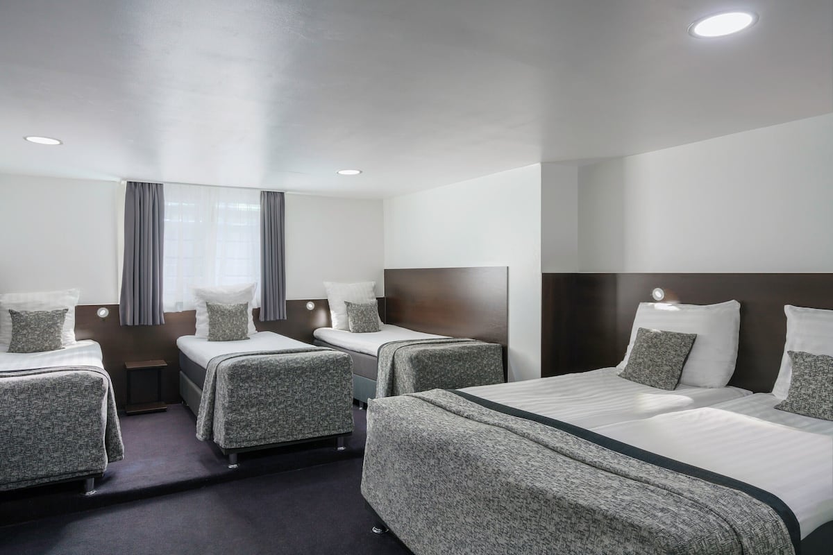 Ein Hotelzimmer mit vier Betten, die in einer U-Form angeordnet sind, einem kleinen Nachttisch, einem großen Spiegel und einem Fenster mit Vorhängen.