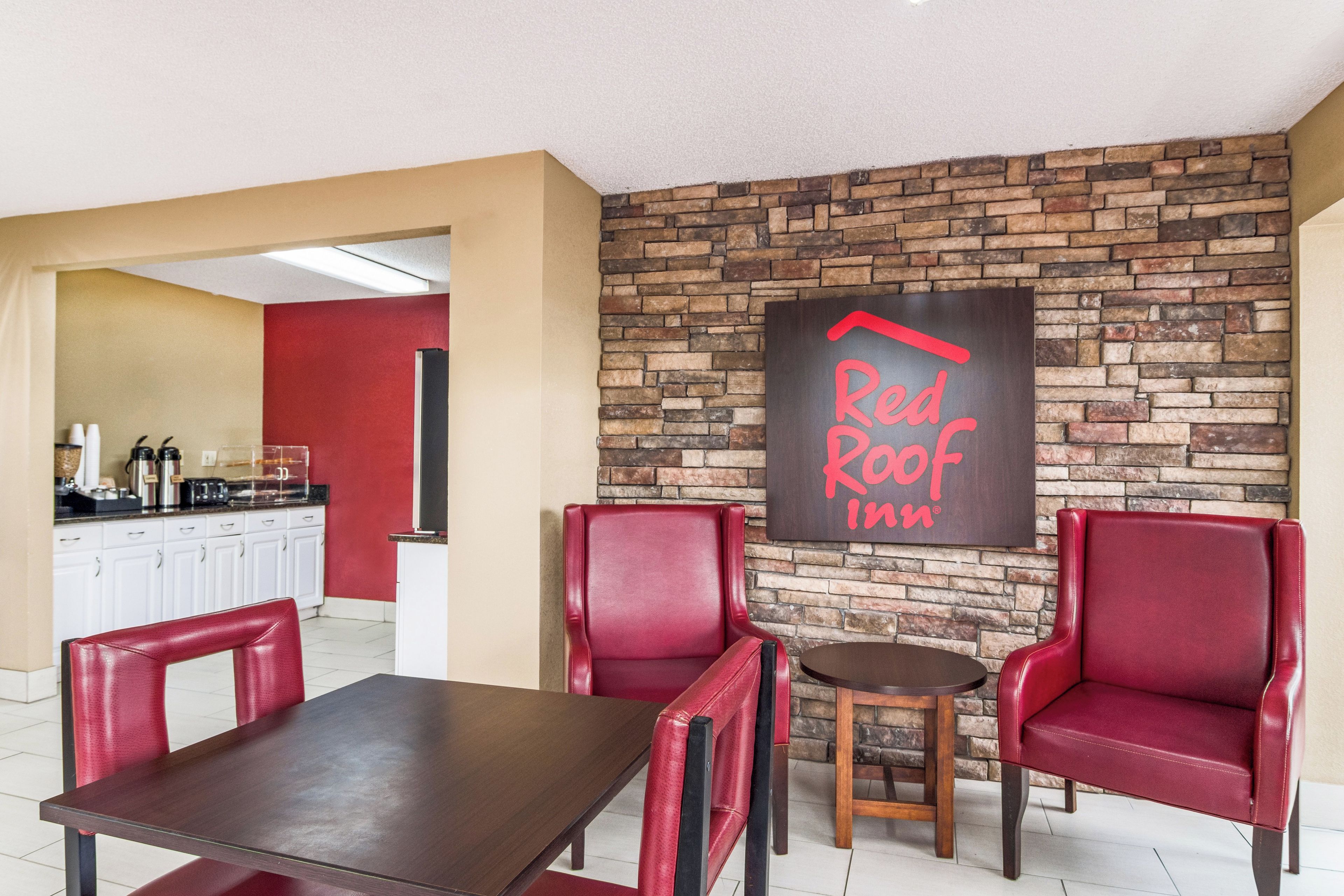 A Red Roof Inn dining area with a brick wall, red leather chairs, a wooden table, and a kitchen in the background.