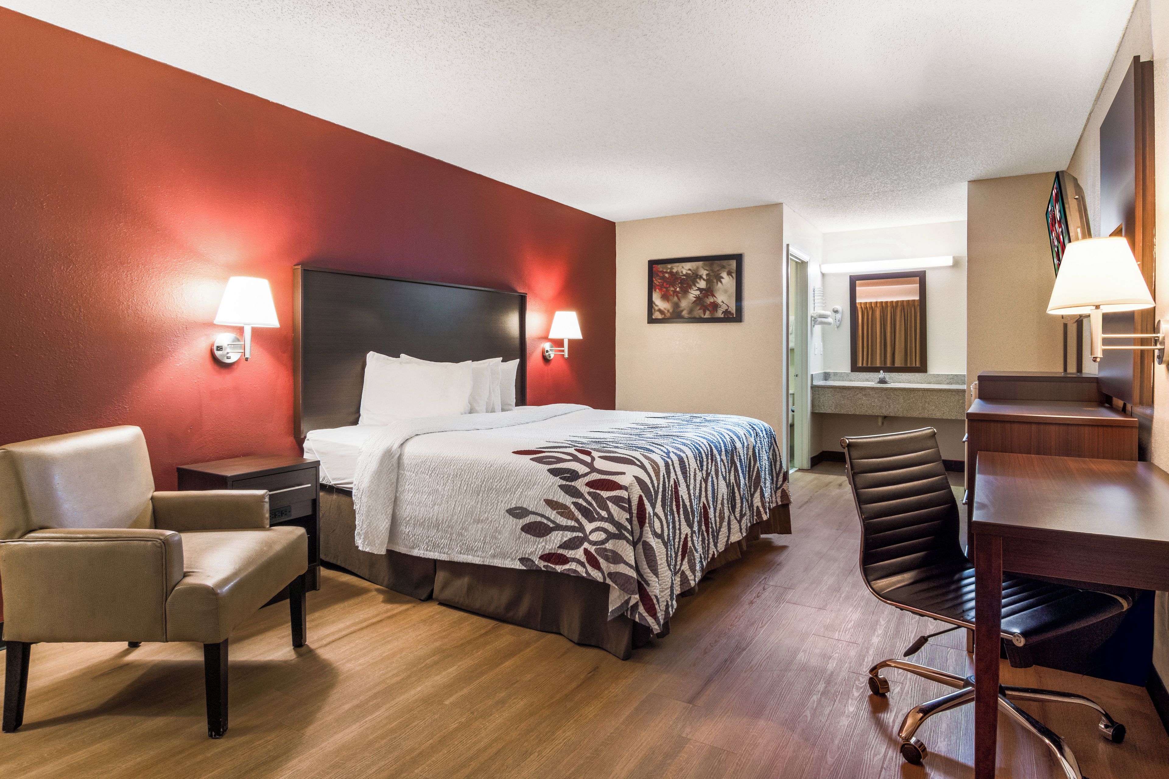 A hotel room with a bed, two chairs, a desk, a mirror, and a bathroom visible in the background.
