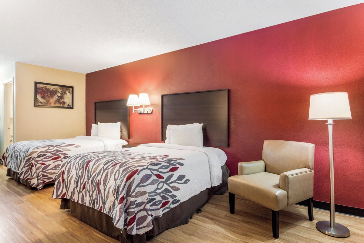 A hotel room with two beds, a chair, a floor lamp, and a painting on the wall.