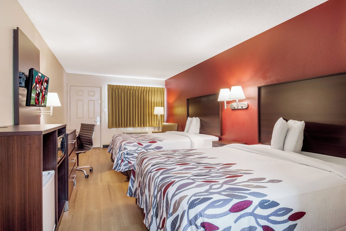 A hotel room with two beds, a desk, a chair, a TV, and a window with curtains.