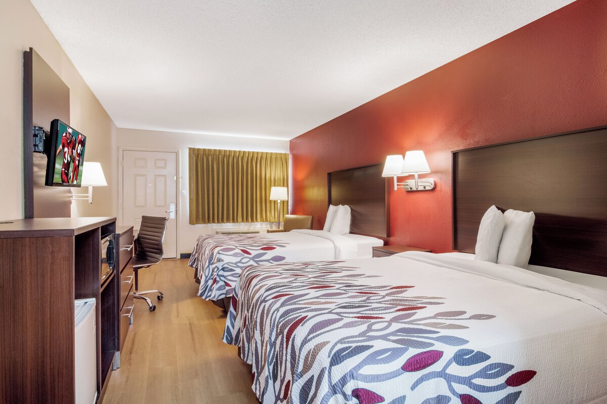 A hotel room with two beds, a desk, a chair, a TV, and a lamp.