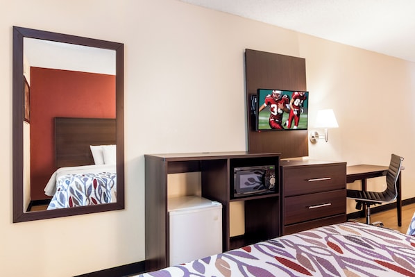 Una habitación de hotel con una cama, un escritorio con silla, un televisor, un microondas y un espejo.