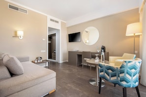 A modern hotel room with a sofa, a dining table, a TV, and a desk.