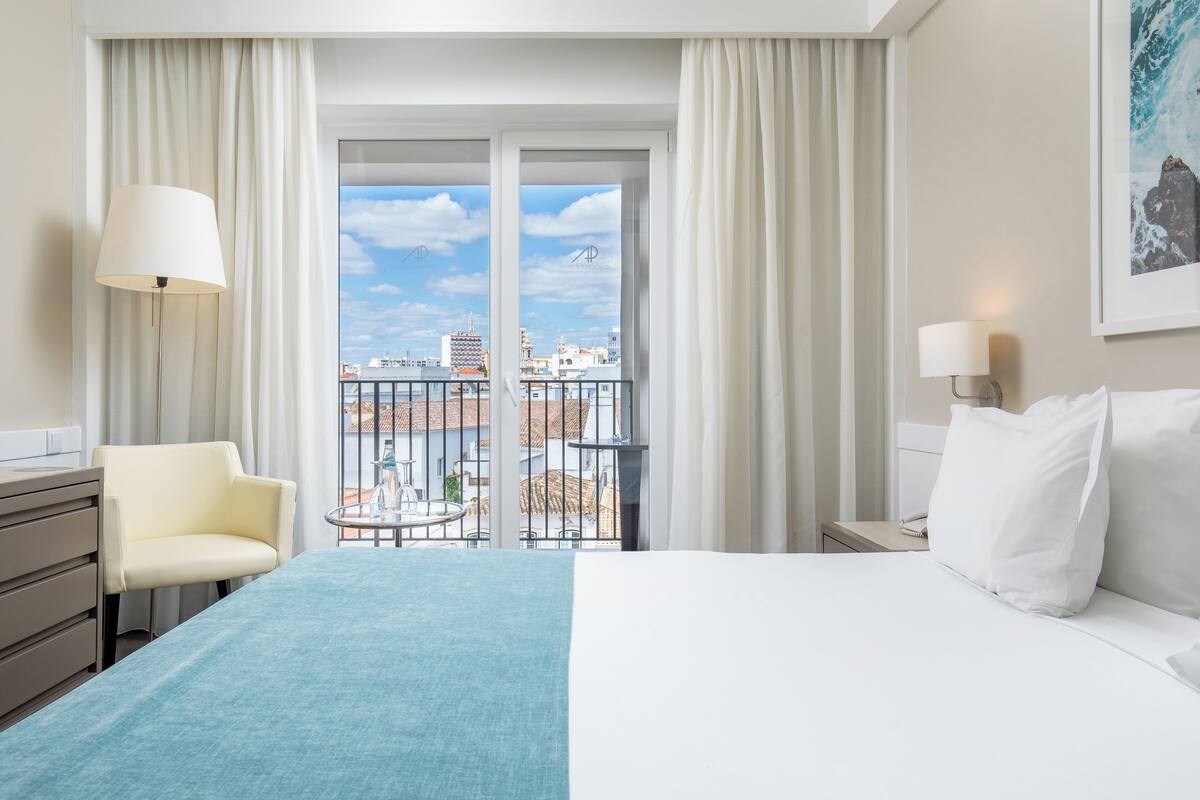 Una camera d'albergo con un letto, una sedia, un tavolino e un balcone con vista sulla città.