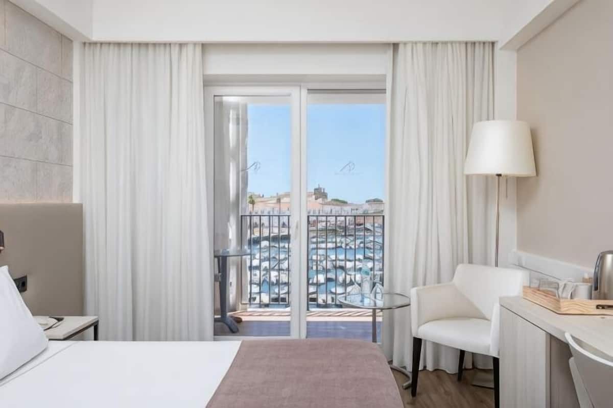 Una camera d'albergo con un letto, una scrivania, una sedia, una lampada e vista su una piscina e degli edifici.