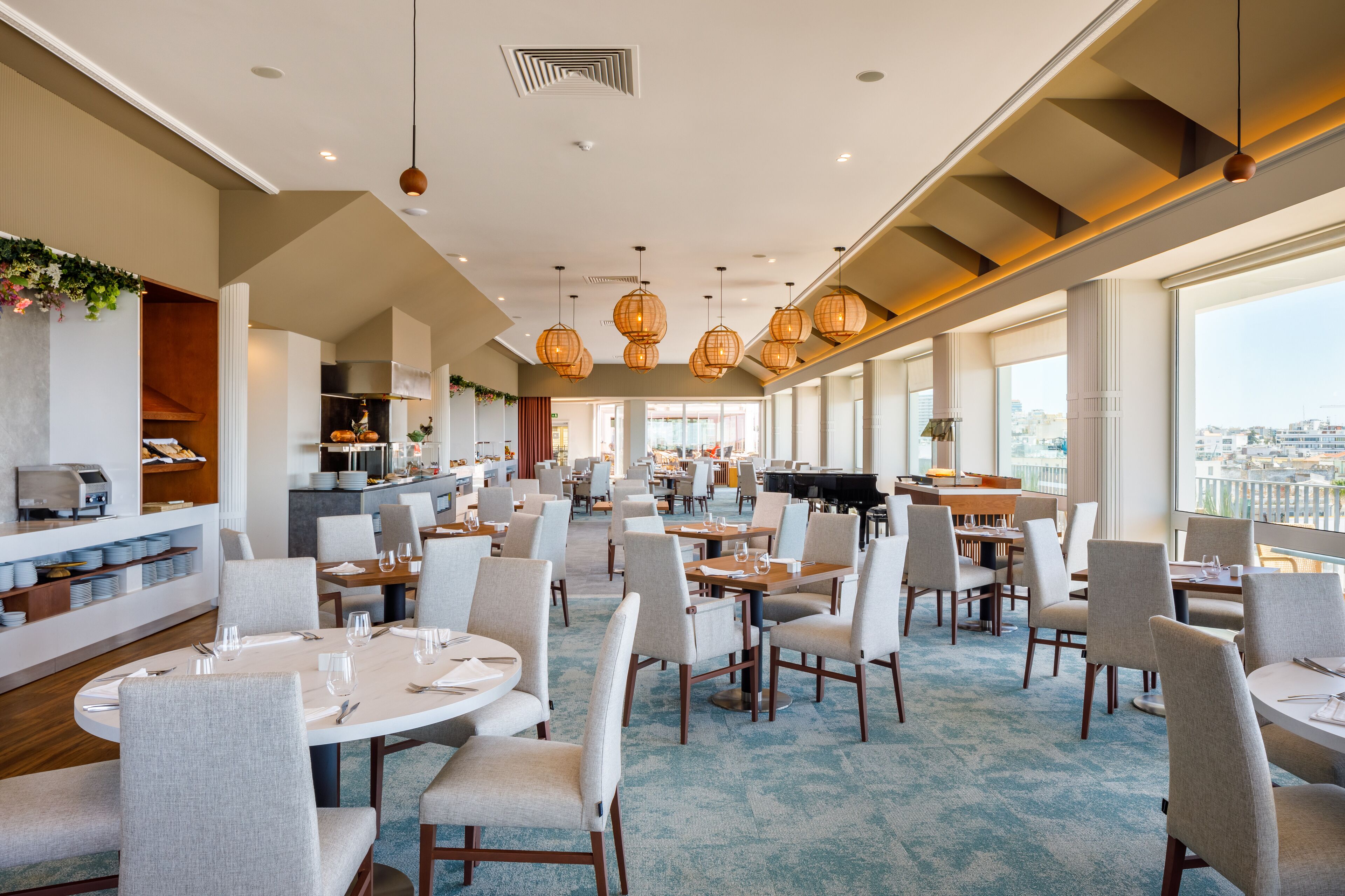 A spacious dining area with round tables and chairs, a kitchen in the background, and large windows offering a city view.