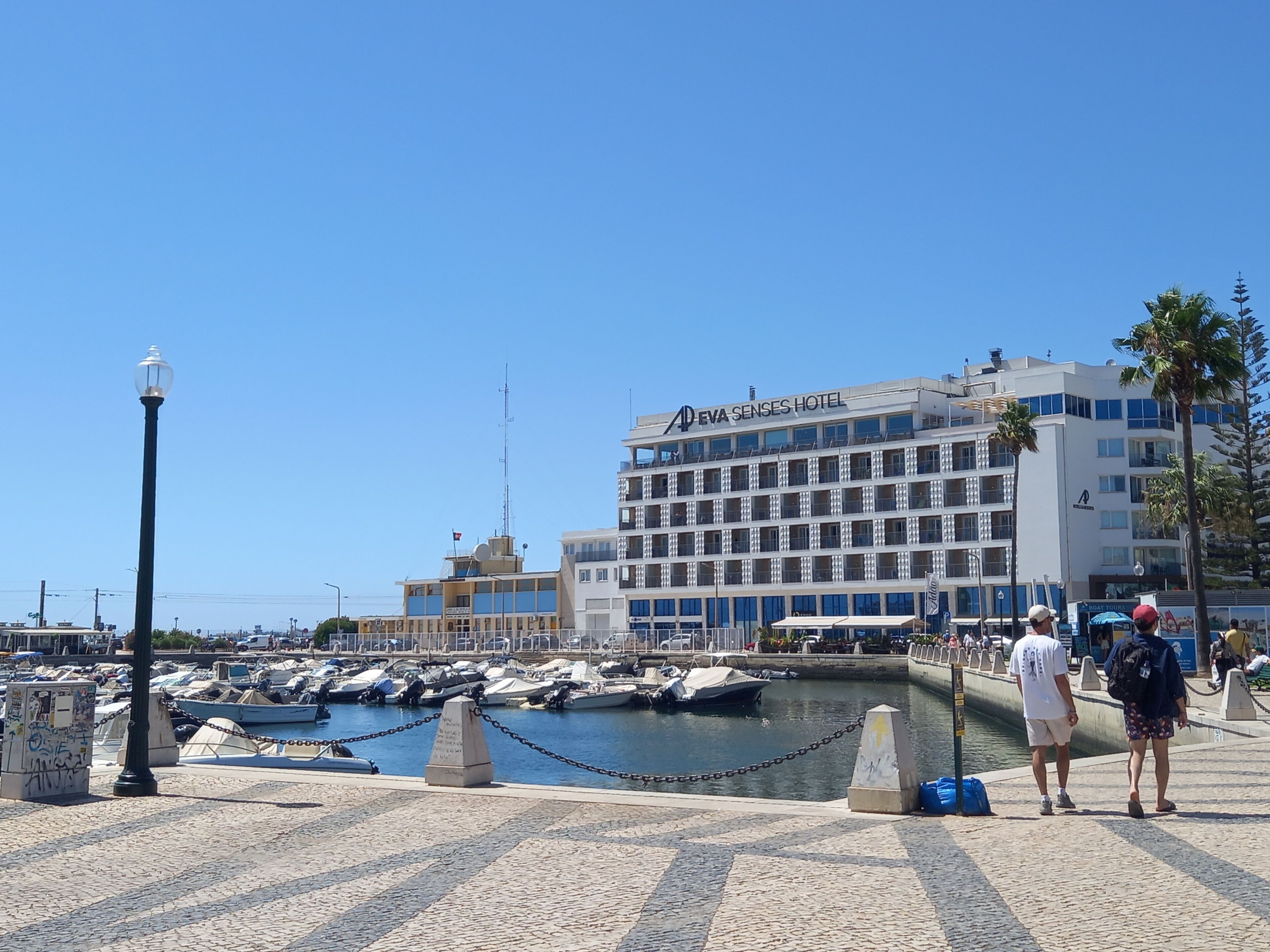 Uma marina com barcos, um hotel chamado 'EVA SENSES HOTEL' e um céu azul límpido.