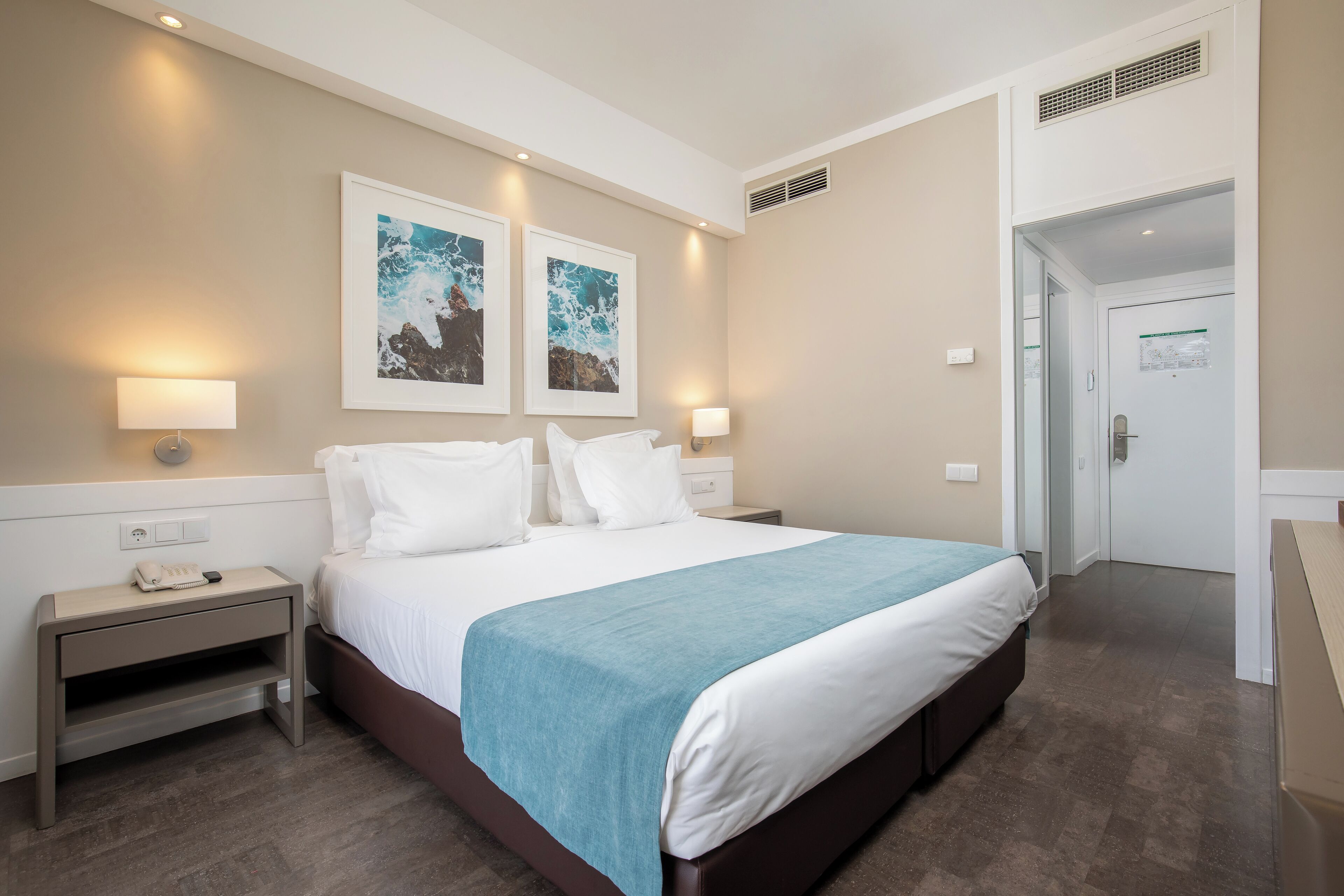 A neatly arranged hotel room with a large bed, two bedside lamps, a nightstand, and two framed pictures on the wall.