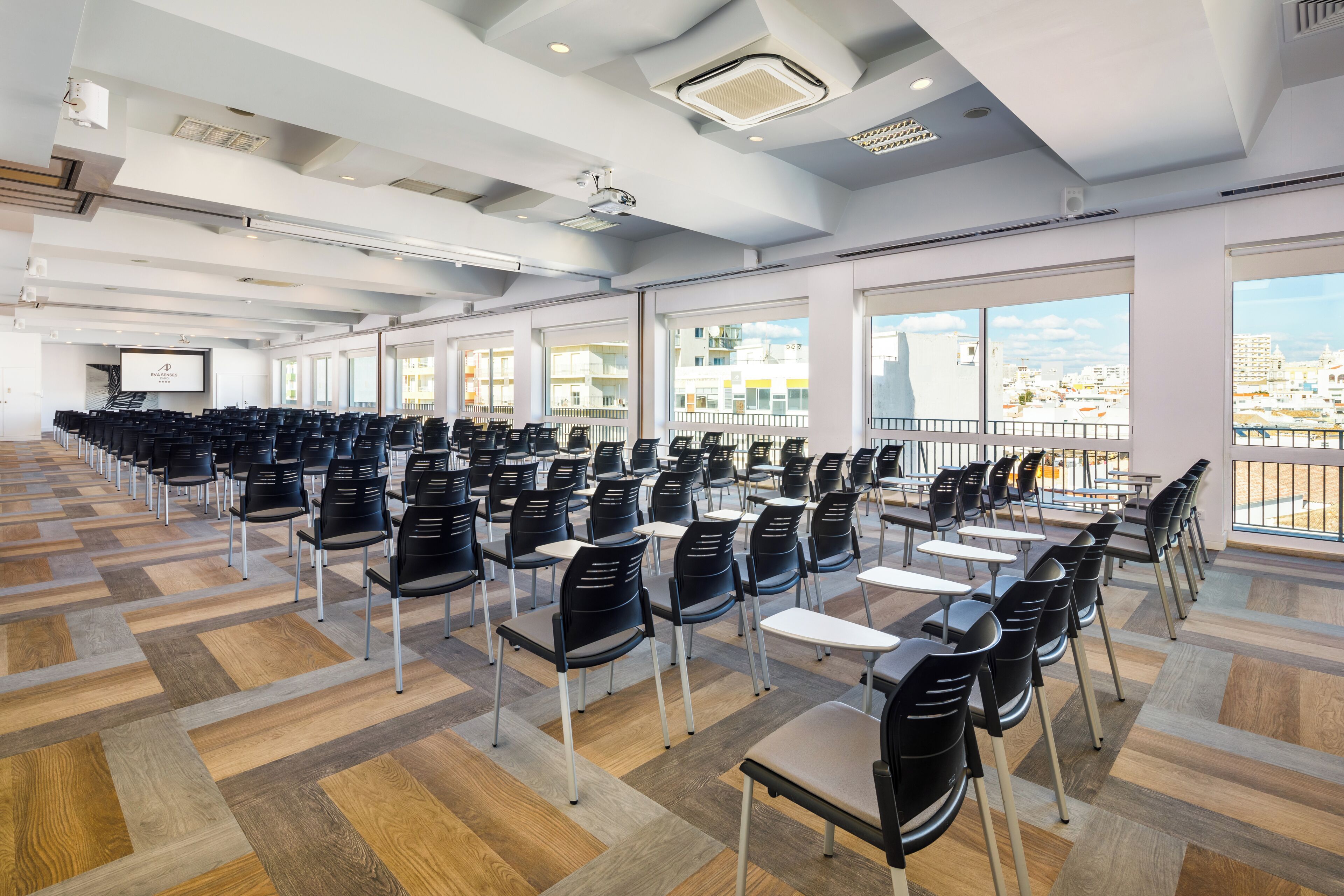 Una amplia sala de conferencias con filas de sillas y mesas, ventanas grandes con vistas a la ciudad y un diseño moderno en el techo.