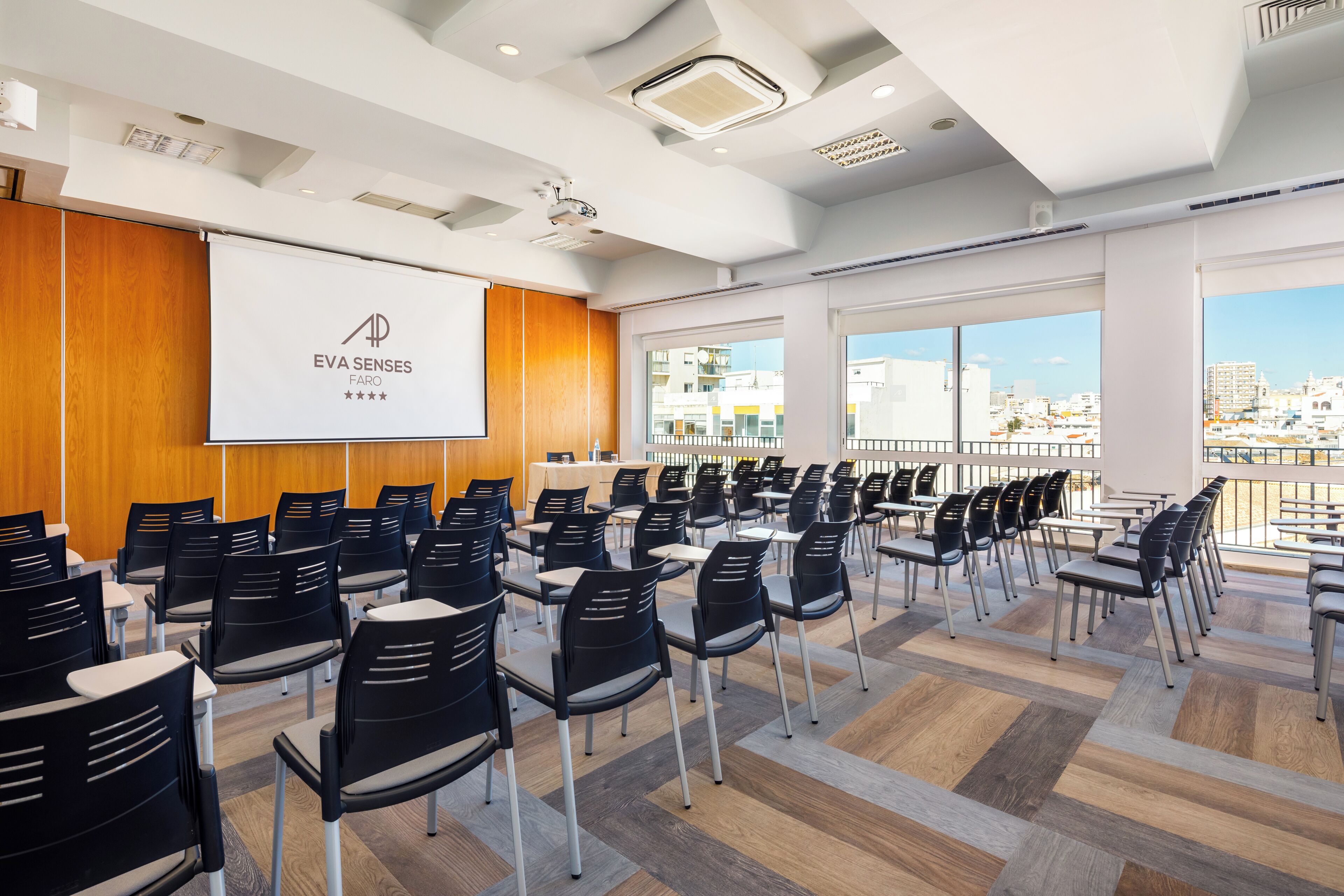 Un salón de conferencias con filas de sillas, una pantalla de proyección y amplios ventanales con vistas a la ciudad.