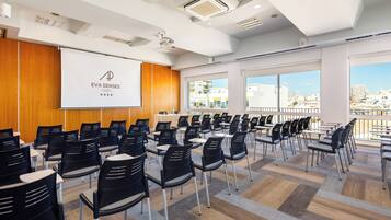 Un salón de conferencias con filas de sillas, una pantalla de proyección y amplios ventanales con vistas a la ciudad.