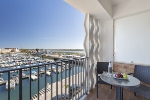 Un balcón con vistas a un puerto deportivo, una mesa con una botella y fruta, y una silla de mimbre.