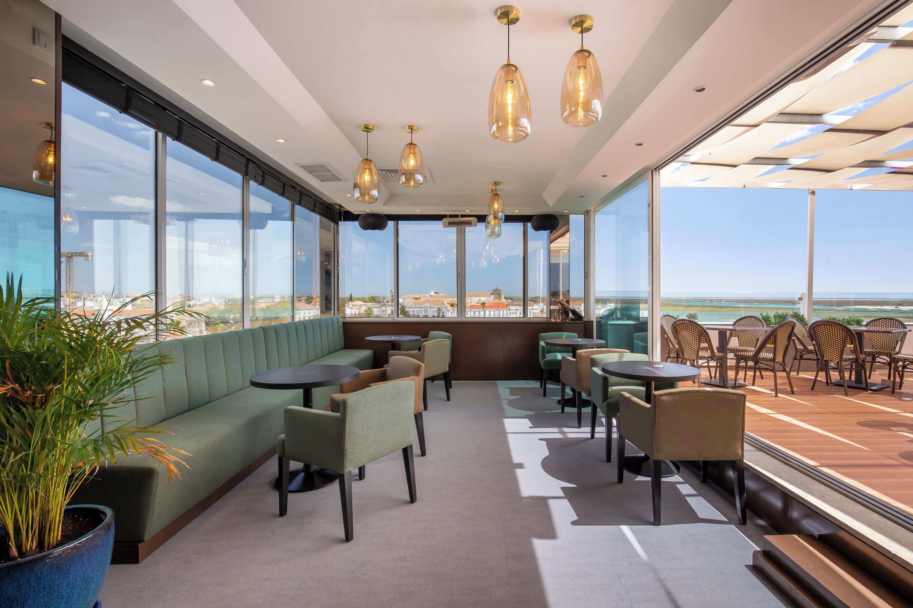 Een modern zithoekje binnen met uitzicht op de oceaan, met een groene bank, meerdere tafels en stoelen.