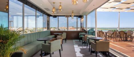 A modern indoor seating area with a view of the ocean, featuring a green sofa, multiple tables, and chairs.