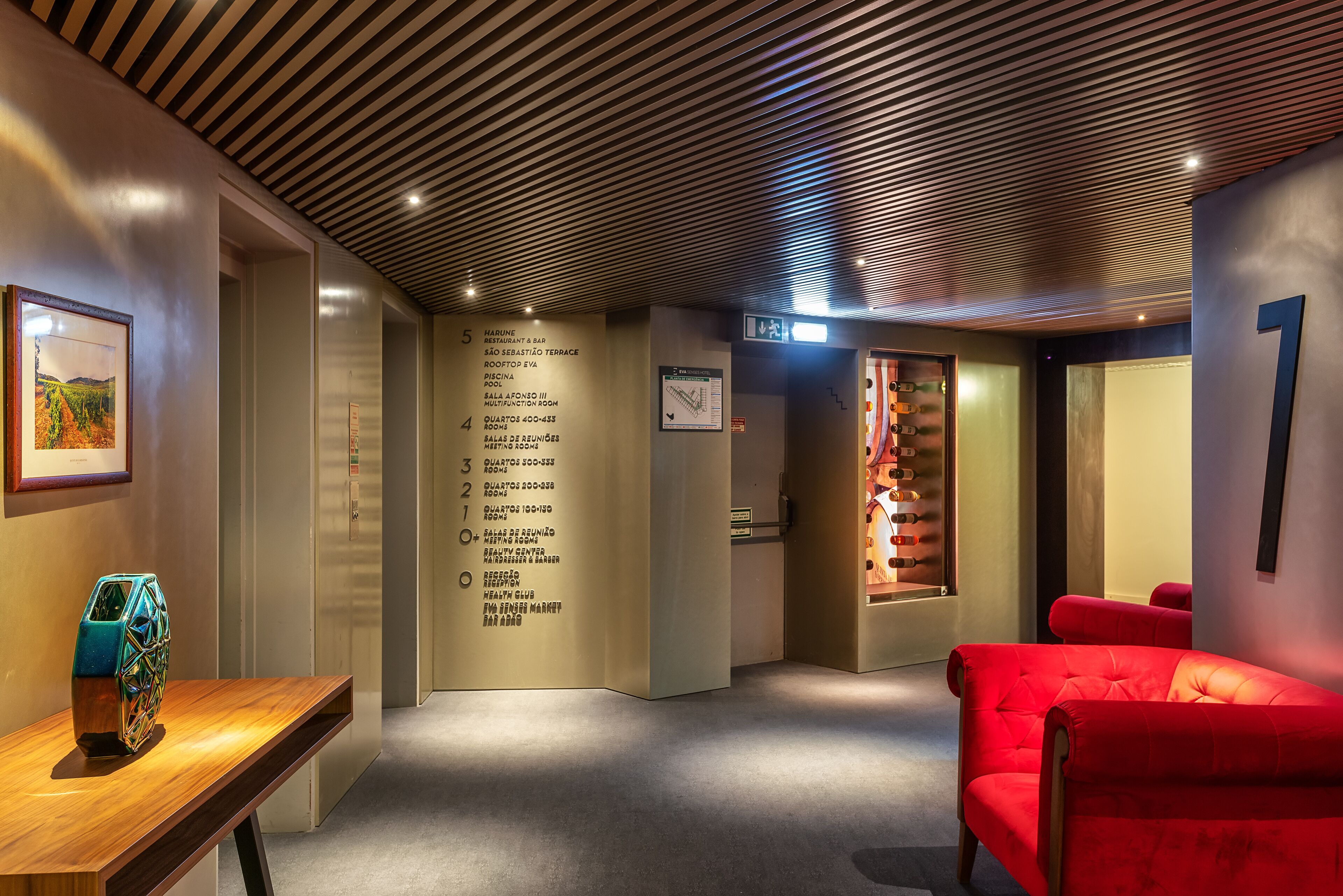 A hotel lobby with a wooden ceiling, a red sofa, a wooden table with a decorative vase, and a wall with room numbers and names.