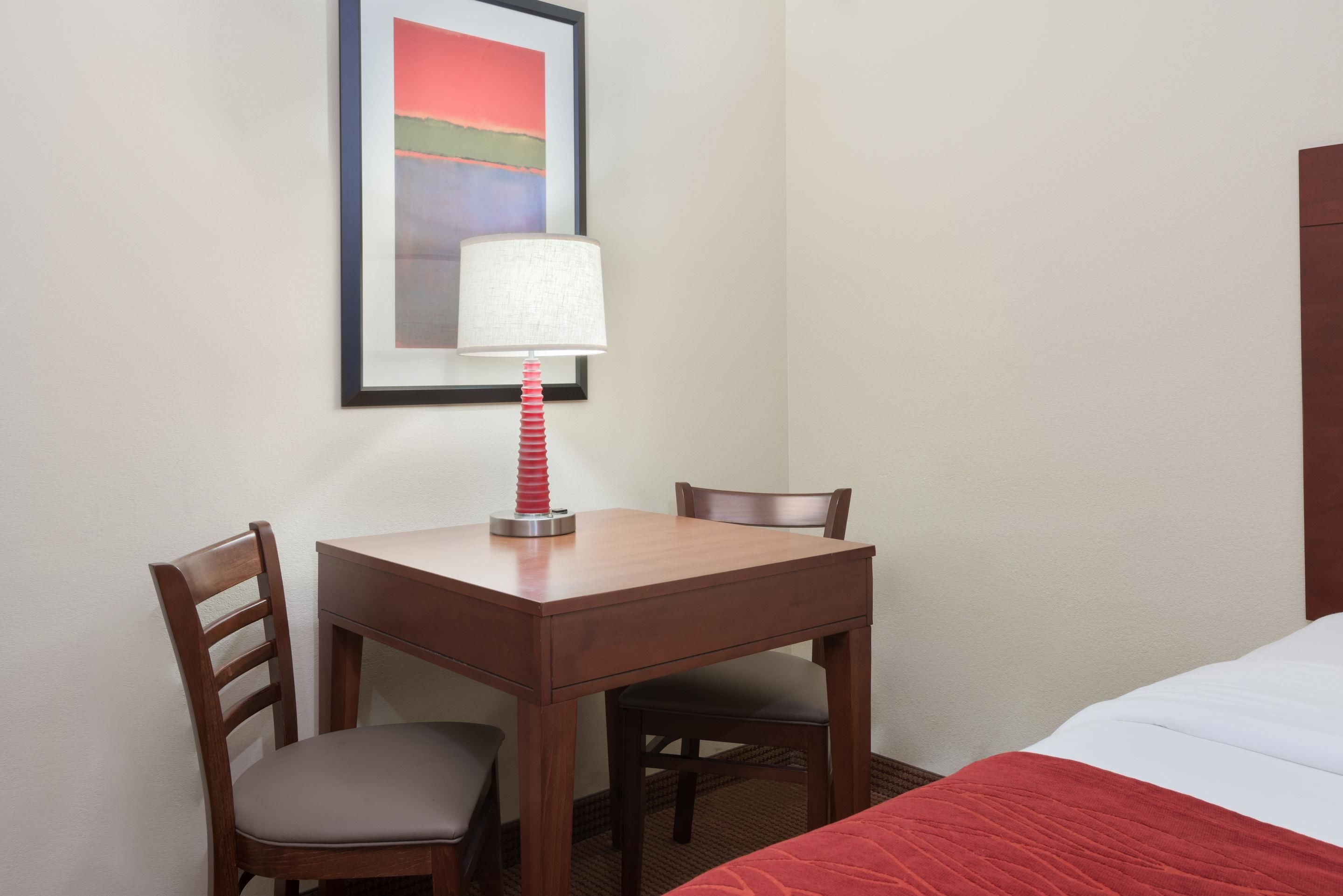 A hotel room with a wooden desk, two chairs, a lamp, a bed with a red blanket, and a framed picture on the wall.