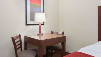 A hotel room with a wooden desk, two chairs, a lamp, a bed with a red blanket, and a framed picture on the wall.