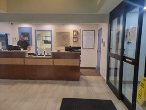 A reception desk with a man standing behind it, a glass door to the right, and a wall-mounted sign above the desk. - Comfort Inn Fayetteville near Fort Bragg (Fayetteville)
