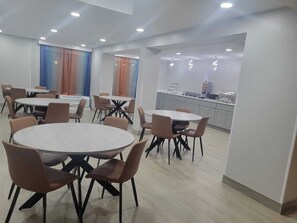 A modern dining area with round tables and chairs, a kitchen area with appliances, and a large window with curtains. - Comfort Inn Fayetteville near Fort Bragg (Fayetteville)