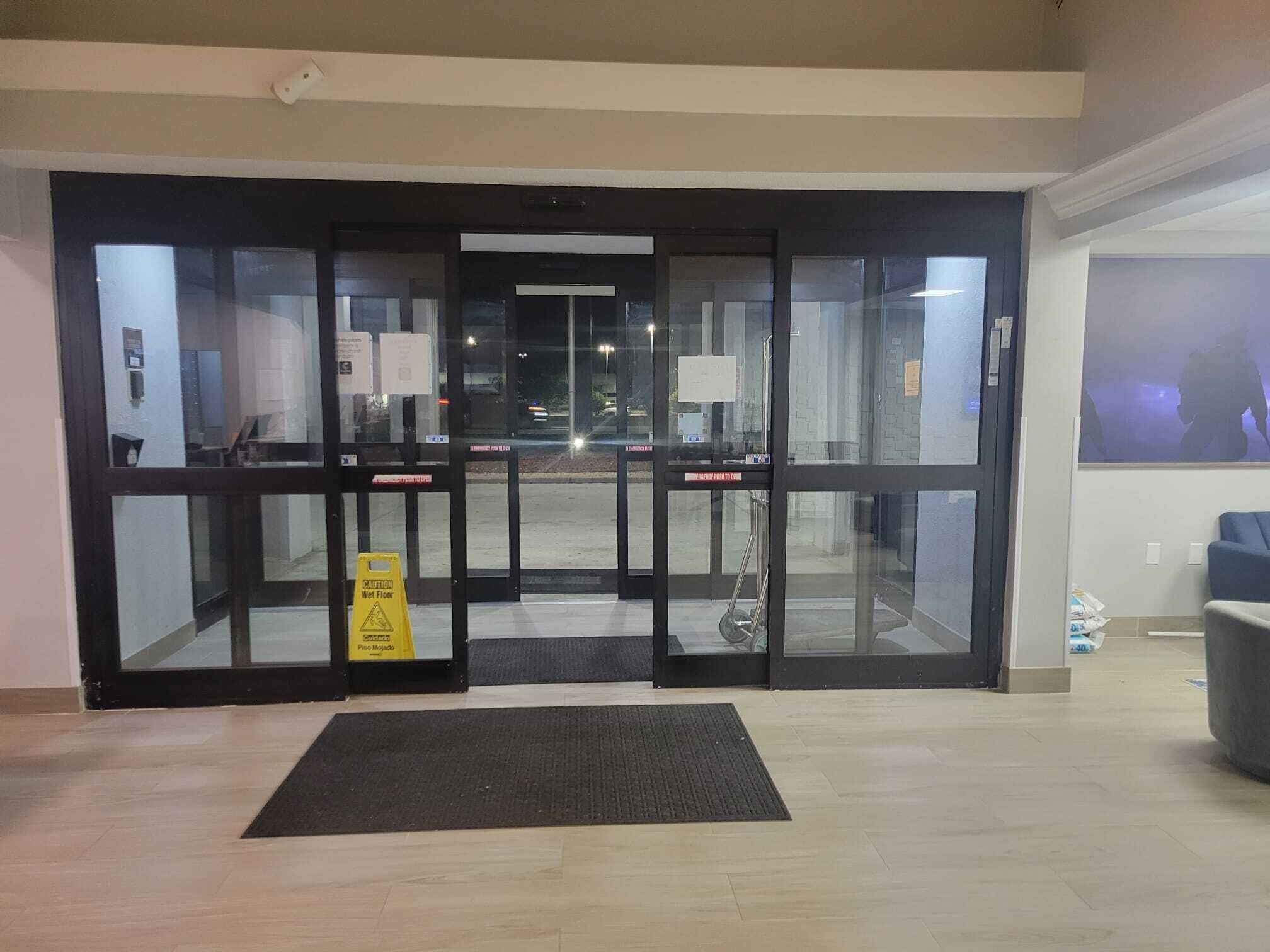 Entrada de hotel com aviso de chão molhado, um tapete e vista para o exterior.