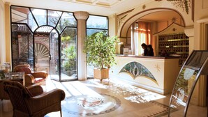 Un vestíbulo con mostrador de recepción, zona de estar, ventanas amplias y una planta decorativa.