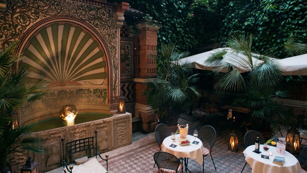 Un patio al aire libre con muros de piedra ornamentados, una fuente y mesas dispuestas para comer.